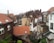 A residential area with closely packed buildings featuring various architectural styles. The rooftops are predominantly covered with red and gray tiles, and some have solar panels. Several chimneys and brick structures are visible, along with bare winter trees and a cloudy sky.
