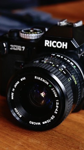 A vintage Ricoh XR7 camera featuring a Rikenon P Zoom lens with specifications of 1:3.4-4.5 and 35-70mm Macro. The camera is black, with a metallic lens and dials, resting on a wooden surface.