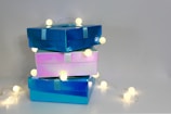 Three glossy gift boxes stacked on top of each other with string lights wrapped around them, casting a warm glow. The boxes are in shades of blue and pink and are tied with ribbons.