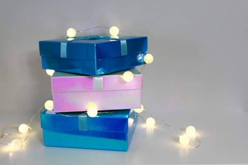 Three glossy gift boxes stacked on top of each other with string lights wrapped around them, casting a warm glow. The boxes are in shades of blue and pink and are tied with ribbons.