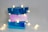 Three glossy gift boxes stacked on top of each other with string lights wrapped around them, casting a warm glow. The boxes are in shades of blue and pink and are tied with ribbons.