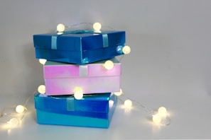 Three glossy gift boxes stacked on top of each other with string lights wrapped around them, casting a warm glow. The boxes are in shades of blue and pink and are tied with ribbons.