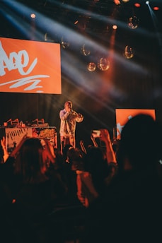 A performer stands on stage with bright red lighting and screens behind them, engaging with a lively audience. The crowd is raising their hands and capturing the moment on their phones. Disco balls and stage lights enhance the energetic atmosphere.