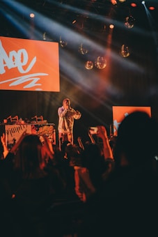 A performer stands on stage with bright red lighting and screens behind them, engaging with a lively audience. The crowd is raising their hands and capturing the moment on their phones. Disco balls and stage lights enhance the energetic atmosphere.