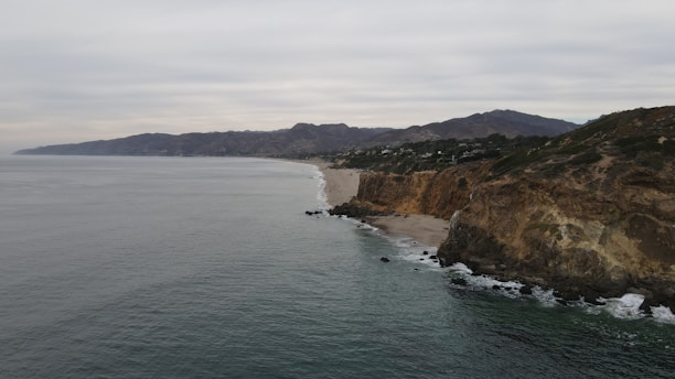 A coastal landscape featuring a long stretch of beach bordered by rugged cliffs on the right. The ocean is calm, with gentle waves meeting the sandy shore. In the background, there are rolling hills under a cloudy sky.