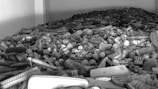 A large pile of various types of brushes and combs is scattered across the surface. The collection includes brushes with different shapes and sizes, some with wooden handles and bristles that are tightly packed. The scene conveys a sense of chaos and neglect reflected in the disorganized and overwhelming accumulation.