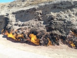 A barren hillside with patches of burning ground, emitting flames and smoke. The flames emerge lengthwise from the earth, suggesting underground combustion. The terrain is dry and rocky, with sparse vegetation on top.