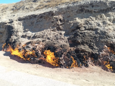 A barren hillside with patches of burning ground, emitting flames and smoke. The flames emerge lengthwise from the earth, suggesting underground combustion. The terrain is dry and rocky, with sparse vegetation on top.