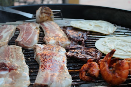 A barbecue grill cooking several items including strips of marinated pork, grilled shrimp, slices of beef, and tortillas. The grill appears to be outdoors, with some cooked and raw items, adding variety to the spread.