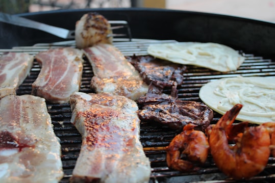 A barbecue grill cooking several items including strips of marinated pork, grilled shrimp, slices of beef, and tortillas. The grill appears to be outdoors, with some cooked and raw items, adding variety to the spread.