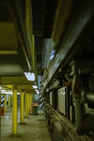 A narrow industrial space featuring a series of yellow support columns leading into the distance. On the right side, there is a complex arrangement of mechanical components and machinery. The lighting is dim and the floor is concrete, giving the area a utilitarian feel. In the background, there is a hint of natural light from a small window.