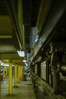 A narrow industrial space featuring a series of yellow support columns leading into the distance. On the right side, there is a complex arrangement of mechanical components and machinery. The lighting is dim and the floor is concrete, giving the area a utilitarian feel. In the background, there is a hint of natural light from a small window.