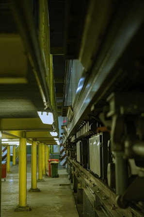 A narrow industrial space featuring a series of yellow support columns leading into the distance. On the right side, there is a complex arrangement of mechanical components and machinery. The lighting is dim and the floor is concrete, giving the area a utilitarian feel. In the background, there is a hint of natural light from a small window.