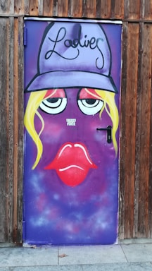 A wooden door painted with a face featuring large eyes, red lips, and blonde hair. The background is primarily purple, with the word 'Ladies' written in an artistic style above the face. A cap or hat element crowns the face, and the door handle is on the right side.