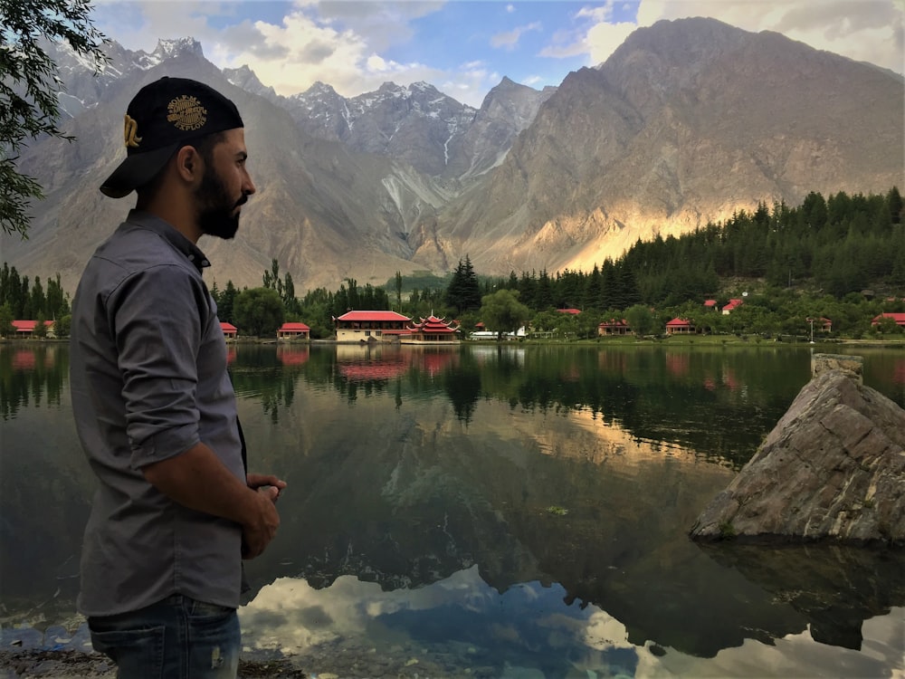 uomo in felpa con cappuccio grigia in piedi vicino al lago e alle montagne durante il giorno