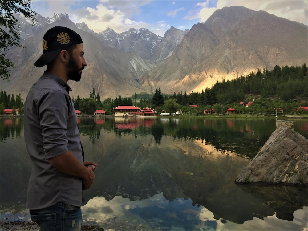 man in gray hoodie standing near lake and mountains during daytime