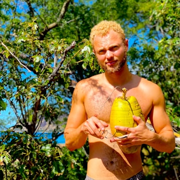 A shirtless man with short curly blond hair is standing outdoors holding a large yellow coconut-like fruit. He is surrounded by lush green trees and foliage with hints of blue sky in the background, indicating a tropical setting.