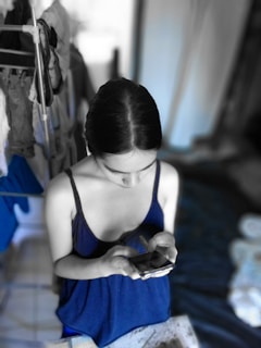 A person with dark hair is looking down at a smartphone while wearing a blue sleeveless top. The background appears to be a domestic setting with clothes hanging on a drying rack and a bed or couch nearby. The surrounding environment is desaturated, highlighting the person and blue elements.