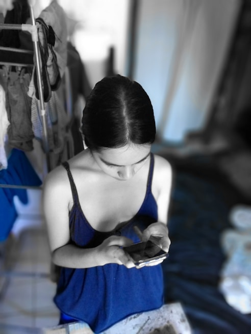 A person with dark hair is looking down at a smartphone while wearing a blue sleeveless top. The background appears to be a domestic setting with clothes hanging on a drying rack and a bed or couch nearby. The surrounding environment is desaturated, highlighting the person and blue elements.