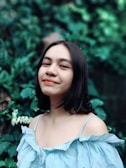 A young woman with long dark hair is wearing a light blue, off-the-shoulder blouse. She is smiling slightly and stands against a backdrop of lush green foliage.