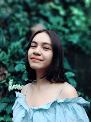 A young woman with long dark hair is wearing a light blue, off-the-shoulder blouse. She is smiling slightly and stands against a backdrop of lush green foliage.