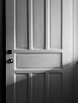 A wooden door with a classic panel design featuring clear vertical and horizontal sections. The image is in black and white, casting soft shadows across the surface.