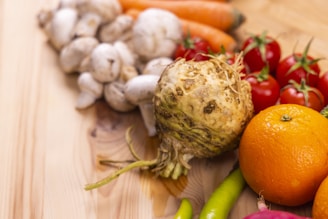 A collection of fresh vegetables and fruits arranged on a wooden surface. The assortment includes mushrooms, carrots, cherry tomatoes, an orange, and a celeriac root, along with chili peppers displayed prominently. The variety showcases different textures and colors, highlighting natural produce.