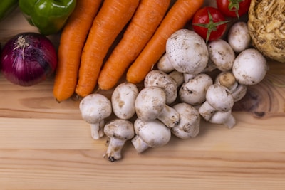 A variety of fresh vegetables are displayed on a light wooden surface. The collection includes white mushrooms, carrots, a red onion, green bell pepper, red tomatoes, and a celery root. The vegetables are arranged in a visually appealing manner, highlighting their natural colors and textures.