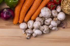 A variety of fresh vegetables are displayed on a light wooden surface. The collection includes white mushrooms, carrots, a red onion, green bell pepper, red tomatoes, and a celery root. The vegetables are arranged in a visually appealing manner, highlighting their natural colors and textures.