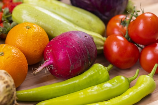A variety of fresh vegetables and fruits are arranged on a wooden surface. Among them are tomatoes, oranges, green peppers, a purple radish, cucumbers, and some leafy greens.