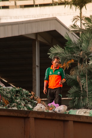 A man in an orange and green uniform stands amidst a pile of plant debris and waste, suggesting a waste management or recycling activity. Large industrial buildings and palm trees are visible in the background.