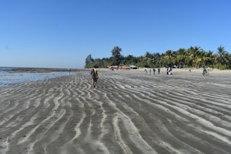 A sandy beach stretches out, forming ripples leading towards the shoreline. Several people walk, some ride bicycles, and a group gathers in the distance under palm trees. The sky is clear and blue, reflecting the calm and sunny atmosphere.