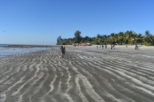 A sandy beach stretches out, forming ripples leading towards the shoreline. Several people walk, some ride bicycles, and a group gathers in the distance under palm trees. The sky is clear and blue, reflecting the calm and sunny atmosphere.