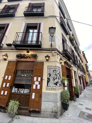 A corner building with multiple balconies featuring dark metal railings. The facade is cream-colored with wooden doors and windows. A large decorative sign reads 'La Cava de Jalan' next to a graphic depicting a person in traditional attire. The entrance is adorned with potted plants and festive decorations.