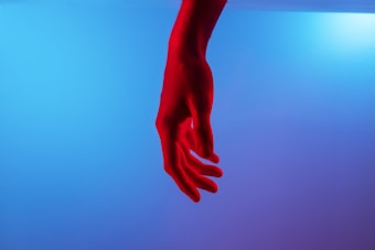 A single hand is prominently illuminated in vivid red against a smooth gradient background transitioning from light blue to purple. The image captures the elegance of the hand in a relaxed and slightly curled position, suggesting an artistic or expressive theme.