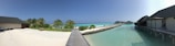 A serene beach scene featuring soft white sand, lush green palm trees, and a clear turquoise ocean. Several bungalows are built on stilts over the water, connected by wooden walkways. The sky is clear and blue, adding to the tranquil atmosphere.