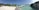 A serene beach scene featuring soft white sand, lush green palm trees, and a clear turquoise ocean. Several bungalows are built on stilts over the water, connected by wooden walkways. The sky is clear and blue, adding to the tranquil atmosphere.