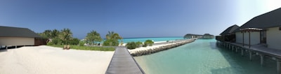 A serene beach scene featuring soft white sand, lush green palm trees, and a clear turquoise ocean. Several bungalows are built on stilts over the water, connected by wooden walkways. The sky is clear and blue, adding to the tranquil atmosphere.