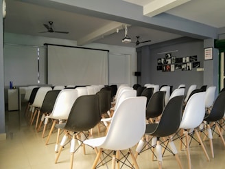 A room with rows of black and white chairs facing a large projector screen. The walls are decorated with framed photographs. Ceiling fans and a projector are mounted above. The room appears to be set up as a small auditorium or meeting space.