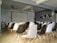A room with rows of black and white chairs facing a large projector screen. The walls are decorated with framed photographs. Ceiling fans and a projector are mounted above. The room appears to be set up as a small auditorium or meeting space.
