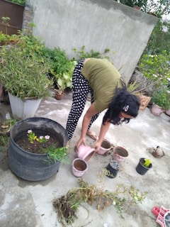 A person is bending over on a concrete surface surrounded by potted plants. They appear to be watering or planting, as they hold a watering can and are positioned above several small pots. Various plants and foliage are visible around, providing a lush and green backdrop.