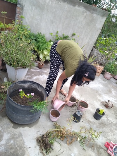 A person is bending over on a concrete surface surrounded by potted plants. They appear to be watering or planting, as they hold a watering can and are positioned above several small pots. Various plants and foliage are visible around, providing a lush and green backdrop.