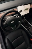 A modern car interior featuring a sleek black steering wheel with a logo, a large touchscreen display showing vehicle information, and a stylish black leather seat. The dashboard has a minimalist design with wooden trim, and the gear selector is visible on the steering column.