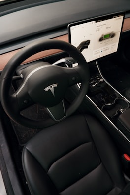 A modern car interior featuring a sleek black steering wheel with a logo, a large touchscreen display showing vehicle information, and a stylish black leather seat. The dashboard has a minimalist design with wooden trim, and the gear selector is visible on the steering column.