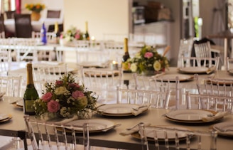 A beautifully arranged banquet hall with elegantly set tables featuring floral centerpieces and champagne bottles. The tables are adorned with white plates and neatly folded napkins. The setting appears ready for a formal event or celebration.