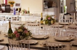 A beautifully arranged banquet hall with elegantly set tables featuring floral centerpieces and champagne bottles. The tables are adorned with white plates and neatly folded napkins. The setting appears ready for a formal event or celebration.
