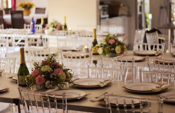 A beautifully arranged banquet hall with elegantly set tables featuring floral centerpieces and champagne bottles. The tables are adorned with white plates and neatly folded napkins. The setting appears ready for a formal event or celebration.