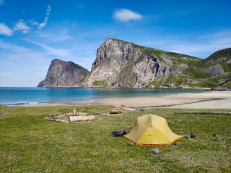 A picturesque coastal landscape features a sandy beach beside a calm blue ocean. Towering cliffs with patches of green grass rise majestically in the background. In the foreground, a yellow tent is pitched on a grassy area near a small arrangement of stones and logs, suggesting a campsite.