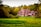 A large red-brick mansion with a complex roof structure and several chimneys is surrounded by lush green grass and dense trees displaying autumn foliage. The architecture features multiple windows and ornate details, enhancing the grandeur of the structure.