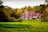 A large red-brick mansion with a complex roof structure and several chimneys is surrounded by lush green grass and dense trees displaying autumn foliage. The architecture features multiple windows and ornate details, enhancing the grandeur of the structure.